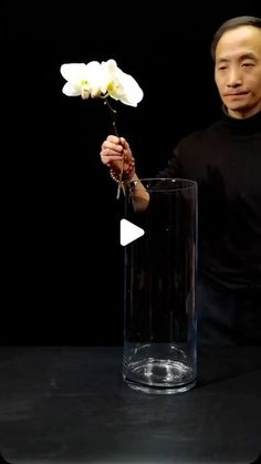 a man holding a white flower in a glass vase with water on the table next to it