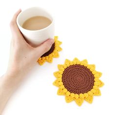 a hand holding a cup of coffee next to a crocheted sunflower coaster