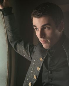 a man in a black jacket leaning against a wall with a large clock behind him