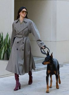 a woman in a trench coat walking her dog on a leash through the city streets