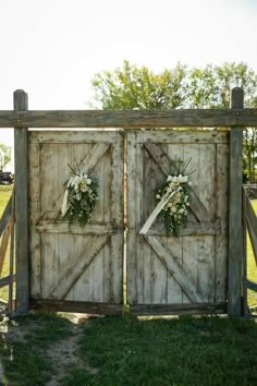 an old wooden gate with two wreaths and flowers on the doors is shown in front of a grassy field