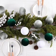 the table is set with silver and green christmas decorations, including pine cones, evergreen branches, berries, white plates and utensils