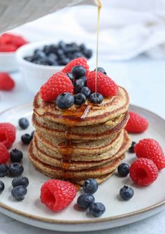 a stack of pancakes with syrup and berries on a white plate next to blueberries and raspberries