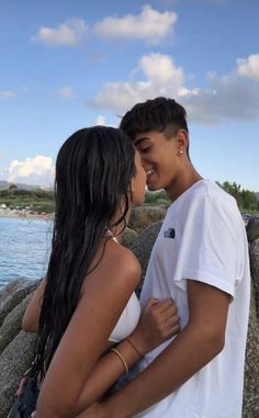 a man and woman standing next to each other near the water with their arms around each other