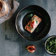 a black plate topped with food next to a bowl filled with greens and fruit on top of a table