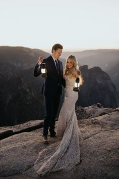 a bride and groom standing on top of a mountain at sunset with their lanterns lit