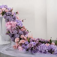 purple and pink flowers are arranged on the floor next to a white vase with greenery