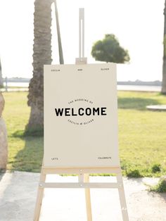 an easel with a welcome sign on it in front of palm trees and grass