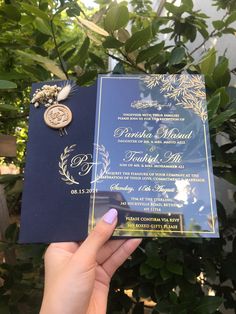 a person holding up a blue and gold wedding card in front of some green leaves