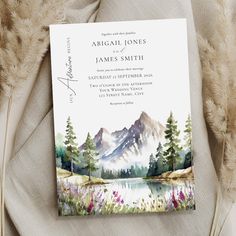 a wedding card with watercolor mountains and flowers on it, sitting next to some feathers