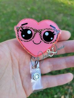 a hand holding a pink heart shaped keychain with sunglasses on it's face