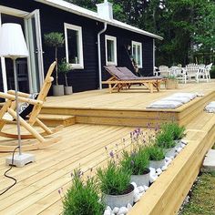 a wooden deck with plants and rocks on the ground next to two lawn chairs in front of a black house