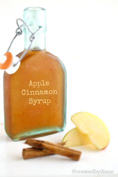 an apple cinnamon syrup in a glass bottle next to sliced apples and cinnamon sticks on a white surface