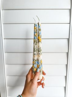 a person holding two candles in front of a white wall with flowers on it and the top one being held up by their hand