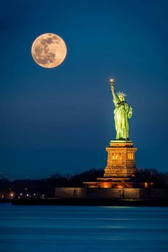 the statue of liberty is lit up at night with the moon in the sky above it