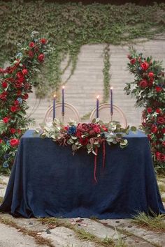 a table with candles and flowers on it in front of an ivy covered wall, surrounded by red roses