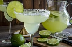 two margarita cocktails garnished with salt and lime on a cutting board next to a pitcher