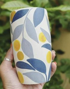 a hand holding a cup with yellow and blue leaves on it in front of a plant