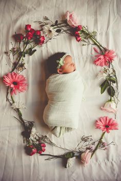 a newborn baby wrapped in a blanket surrounded by pink flowers and greenery on a white sheet