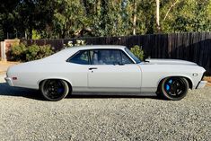 a white muscle car parked on gravel in front of a wooden fence