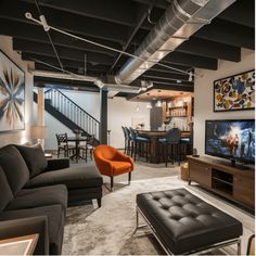 Modern basement living area with a sectional sofa, orange chair, TV, and bar with stools in the background. Chic Basement Ideas, Low Ceiling Basement Ideas, Ceiling Basement Ideas, Chic Basement, Basement Unfinished, Movie Rooms, Ceiling Basement, Low Ceiling Basement, Exposed Ceiling