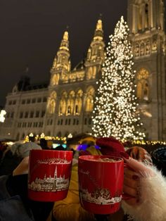 two red coffee mugs in front of a christmas tree with lights on the building