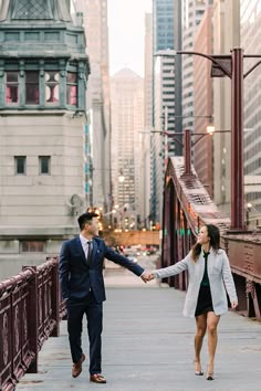 a man and woman holding hands while walking across a bridge in the middle of a city