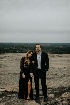 a man and woman standing next to each other on top of a rocky hill with the sky in the background