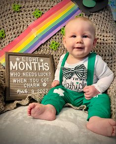 a baby wearing green overalls sitting next to a sign that says eight months who needs luck when you got charm