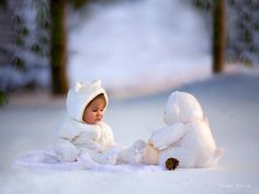 a baby sitting in the snow next to a teddy bear wearing a white coat and holding a stuffed animal