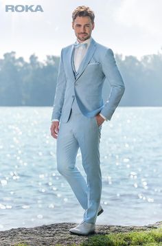 a man in a light blue suit stands on the shore by the water with his hands in his pockets