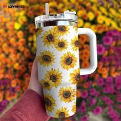 a hand holding a coffee cup with sunflowers on it in front of flowers