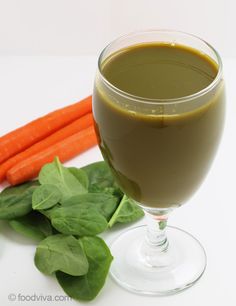 a glass filled with liquid next to carrots and spinach leaves on a white surface