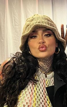 a woman with long black hair wearing a hat and making a peace sign while standing in front of a white curtain