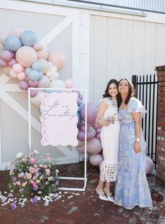 two women standing next to each other in front of a sign with balloons on it