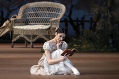 a woman sitting on the ground reading a book in front of a wicker chair