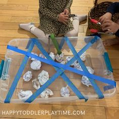 two children sitting on the floor playing with construction paper and scissors in a plastic container