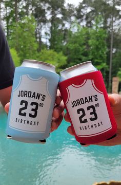 two people holding up cans of soda next to a pool with trees in the background