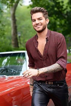 a man with tattoos standing in front of a red car and smiling at the camera