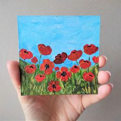 a hand holding up a piece of paper with red flowers painted on it and blue sky in the background