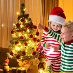 two young children standing next to a christmas tree with lights and ornaments on the table