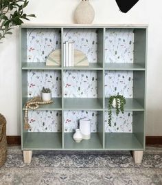 a green bookcase with plants and books on it in front of a white wall