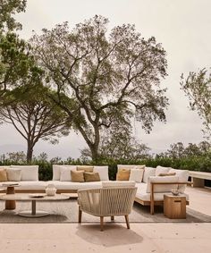 an outdoor seating area with white couches and wooden tables in front of some trees