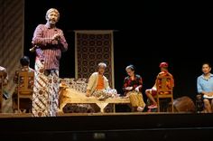 a man standing on top of a stage next to other people sitting in wooden chairs