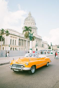 an old yellow car driving down the street in front of a large building with a dome on top