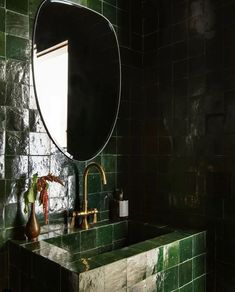 a bathroom with green tiled walls and a round mirror above the sink, along with a vase filled with flowers