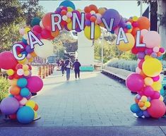 an arch decorated with balloons and the words carnival on it's sides, along with people walking in the background