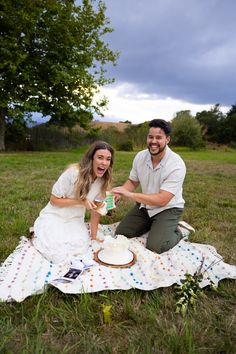 a man and woman sitting on a blanket in the middle of a field eating cake