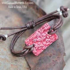 a leather cord bracelet with a red square pendant on it's end, sitting on top of an old rusty piece of metal