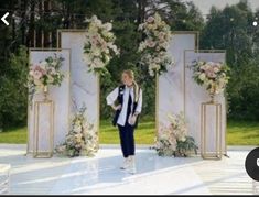 a woman standing in front of an arch with flowers on it and another person walking down the aisle